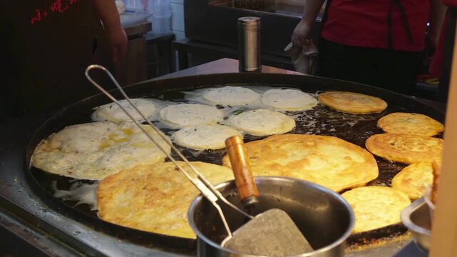 Vendor removing cooked Cong You Bing pancakes from large griddle in Shanghai. Traditional Chinese flatbread preparation and hawker center cooking for local culinary culture