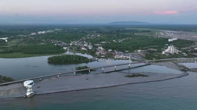 Horizontal drone aerial footage frame capturing the long wooden pedestrian bridge in Anaklia, showcasing the river delta meeting the Black Sea beach, a small green island, and coastal resort