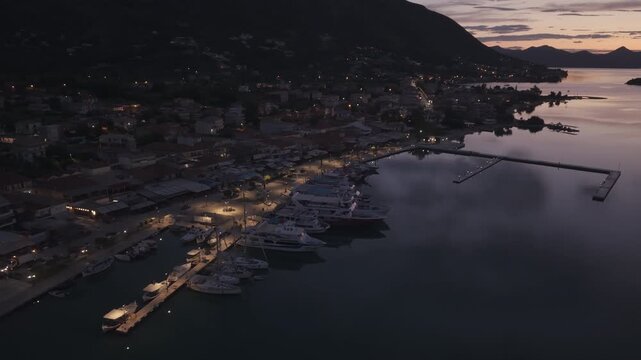 Atmospheric drone footage captured during blue hour above a coastal harbor town. Marina lights begin to glow as yachts rest peacefully along the waterfront, while calm waters reflect the fading night 