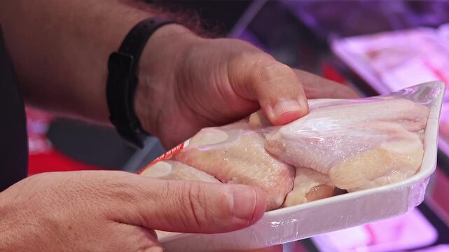 Customer inspecting packaged chicken wings in supermarket meat department, checking fresh poultry product before purchase. Close up consumer selection, refrigerated grocery shopping.