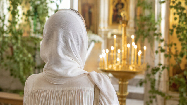 Woman in white headscarf praying in orthodox church during Trinity Sunday. Religious person standing in front of candles and sacred icons. Spiritual meditation and faith practice.