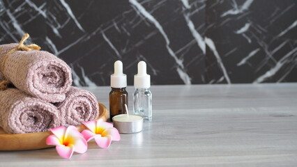 Luxury spa still life with essential oil dropper bottles (amber and clear glass), a scented candle, soft pink mauve towels tied with rope, and pink frangipani flowers on a light gray wooden table. Pre © kanjana