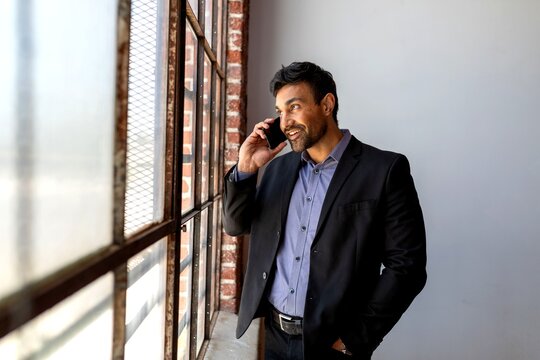 Man in a suit talking on the phone by a large window, smiling.