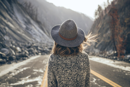 Mountain road landslide person back view empty road rocky slope wind hat sweater daylight woman standing highway canyon road block overexposed sky outdoor travel solitude rear view cliff damaged