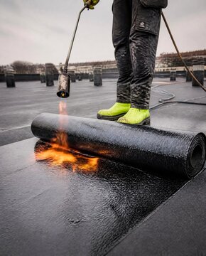 Professional roofer working on flat roof, applying bituminous waterproofing membrane, propane gas torch