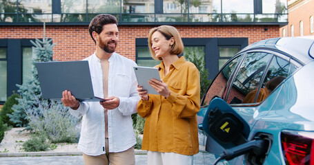 Obraz premium Business man holding a laptop and business woman holding a tablet standing outdoors near a charging electric car, looking at device screens and talking.