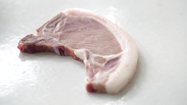 A raw bone-in pork chop placed on a clean white surface, showcasing its light pink meat and white fat layer. This fresh cut of pork is ideal for grilling, frying, roasting