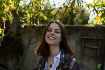 Obraz premium Happy young woman outdoors in a plaid shirt, smiling toward the camera, warm sunlight, casual lifestyle portrait in a leafy garden setting, carefree positive mood