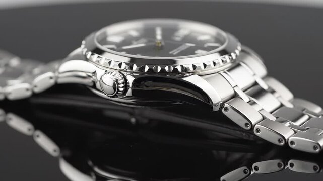 Close-up side profile of a stainless steel wristwatch with a black dial and metal bracelet, reflecting on a glossy surface.
