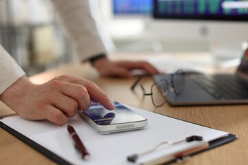 Fototapeta premium Financial trading specialist using smartphone at wooden desk in office, closeup