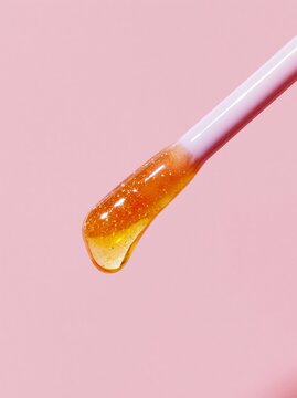 Extreme close-up of lip oil applicator wand with glossy orange liquid against a soft pink background, showcasing the product's texture and shine