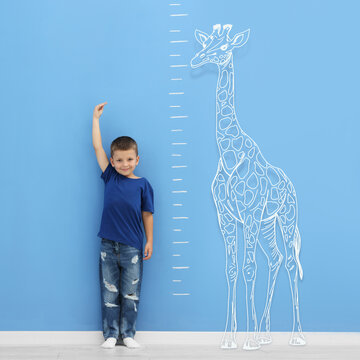 Little boy measuring his height near blue wall with scale and drawing of giraffe