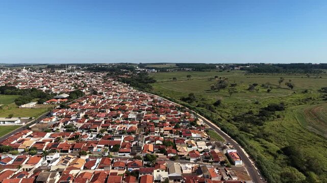 Aerial view of a sprawling residential neighborhood with red tiled roofs bordering a vast green agricultural field under a clear blue sky