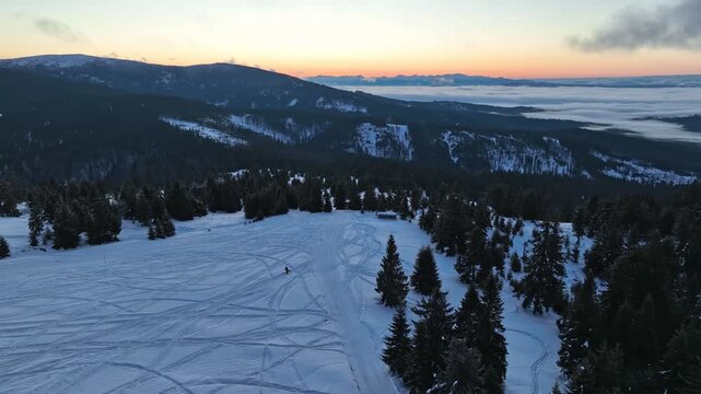 Drone orbital flight around snowboarder silhouette walking on snow covered mountain peak before sunrise with misty valleys