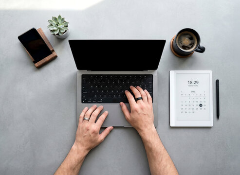 Top down view of male hands typing on a laptop computer at a minimalist concrete desk with an electronic calendar, smartphone, and black coffee.