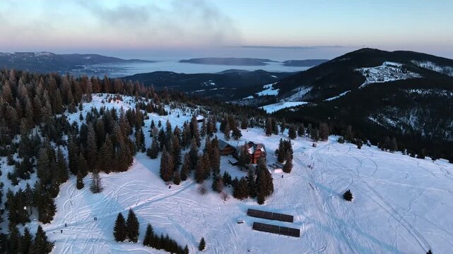 Aerial pull back from traditional wooden mountain chalet at sunrise with tourists silhouettes on snow meadow