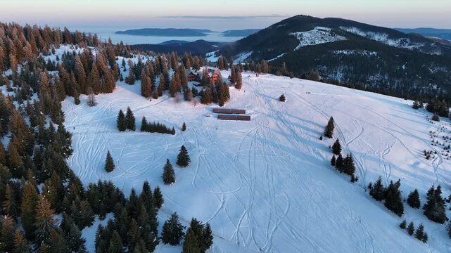 Aerial orbit tracking traditional wooden chalet on snow covered mountain meadow during warm winter sunrise