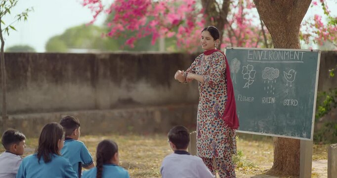 Indian young adult woman do dance teach to girl boy enjoy day under tree rural area teen child group wear uniform sitting on mat look tutor learn basic step moves have fun joy open class field place