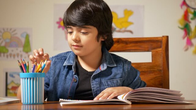 Portrait of a cute Indian boy choosing colored pencil to draw pictures - School homework. Charming little boy sitting on a wooden chair in his study room and completing his holiday homework 
