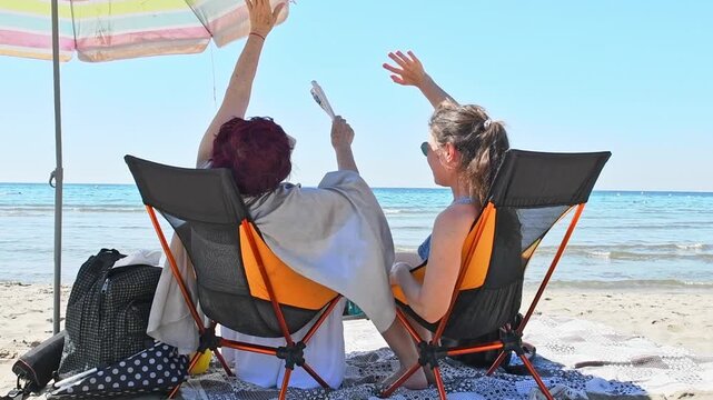 Two women, an older woman and a younger woman, relax in beach chairs under an umbrella, chatting, waving by the sea on a sunny summer day. summer leisure accessories. tourism in a hot country