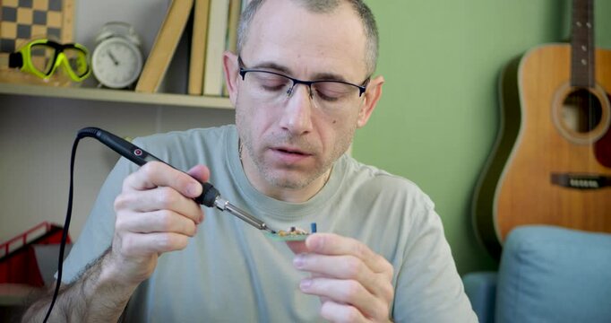 Man solders electronic circuit board while holding it in hand at home workstation. Precision work, technical craftsmanship and attention to detail highlight modern electronics repair.