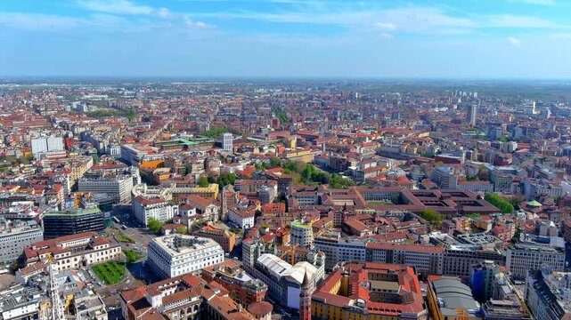 4K aerial drone shot flying over Milan, Italy, capturing the vast urban cityscape, featuring historic terracotta rooftops, modern buildings, and prominent church spires under a blue sky.
