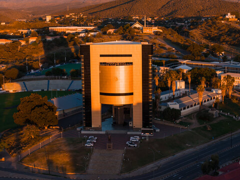 Aerial view of the golden glass Independence Memorial Museum in Windhoek, facing Robert Mugabe Avenue, with Sam Nujoma statue, Alte Feste, stadium, cars, and hills.