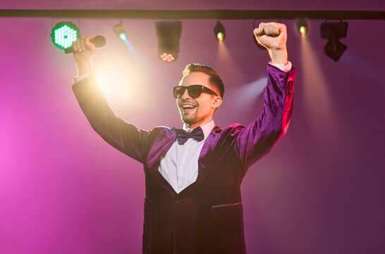 Singer showman presenter man with microphone on stage. Cheerful performer in tuxedo under concert spotlight, arms raised and smiling in vibrant lighting. Winning success and celebration theme.