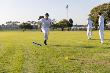 Obraz premium Male teammates wearing white cricket kit sprinting through blue cones on sports field, yellow cone