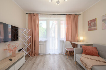 Fototapeta premium cozy living room with a grey sofa, peach curtains, a white desk, and a TV unit; light wood flooring, abstract art, and a folding drying rack add warmth and functionality