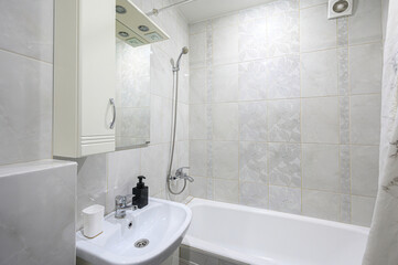 Fototapeta premium compact bathroom with white marble-patterned tiles, a bathtub with a handheld shower, a wall-mounted sink, a mirrored medicine cabinet, and a wall-mounted extractor fan