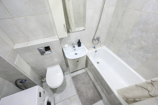 compact bathroom with light marble-patterned tiles, a bathtub with a shower hose, a wall-mounted sink unit, a toilet, and a washing machine tucked in the corner