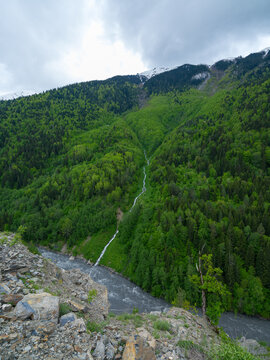 Vertical landscape photograph capturing a deep mountain gorge from a high viewpoint, showcasing lush green alpine pastures, a winding stream, dense forests, and a massive snow-covered mountain range