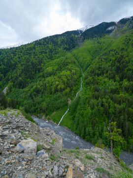 Vertical landscape photograph capturing a deep mountain gorge from a high viewpoint, showcasing lush green alpine pastures, a winding stream, dense forests, and a massive snow-covered mountain range