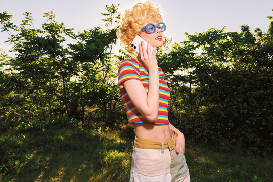 A stylish young woman with curly blonde hair talks on a phone outdoors, wearing a striped crop top and beige skirt, posing confidently amid sunlit greenery in retro film color.