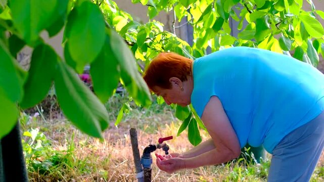 Refreshing herself with cool water from a garden tap a grandmother splashes her face and enjoys simple moment outdoors. The scene reflects rural living personal care summer heat and everyday routines.