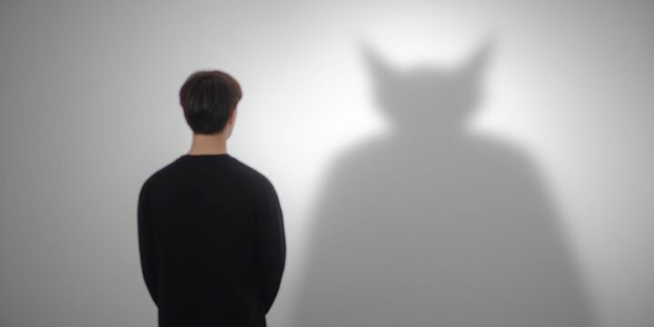 a man stands in front of a wall, with a scary monster's shadow cast on the white wall behind him. the image is monochrome and simple, a work of photography.