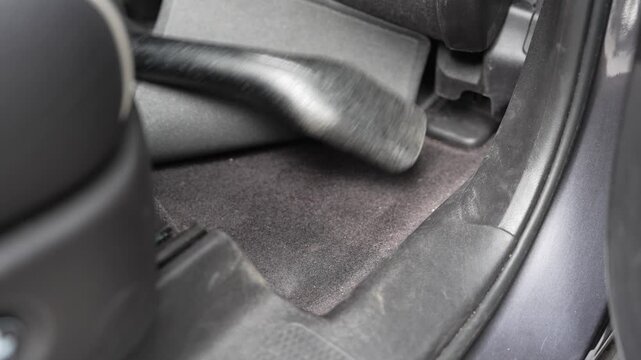 Close-up of a vacuum cleaner nozzle cleaning the carpet floor underneath a lifted mat inside a vehicle. A young helper participating in family chores and automotive interior detailing.