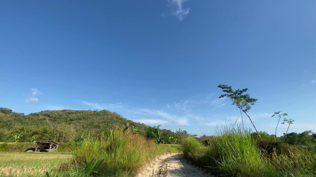Wide shot of a rural dirt path with green hills and clear blue sky