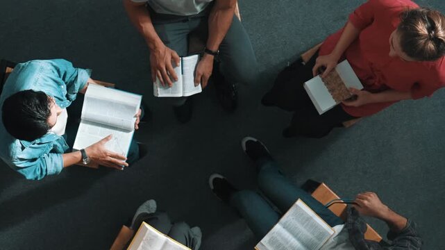 Top down view of prayer reading at bible book and sitting in circle with bible book on laps. Aerial view of diverse people looking at book while studying with faith, trust and hope, calm. Symposium.