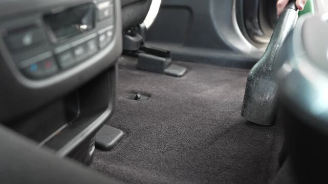 A close-up view of a man's hand holding a vacuum cleaner nozzle and moving it across the dark carpet floor in the rear passenger compartment of a vehicle. The video shows the vacuuming process