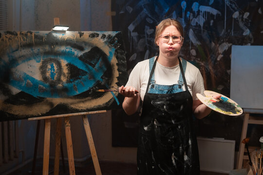 A Caucasian female artist creates a masterpiece in an art studio. A contemporary painting on canvas. Acreative, modern, and successful artist stands looking at the camera, pouting.