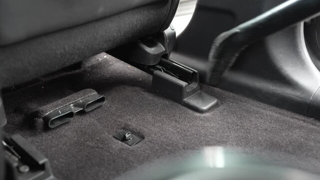 A close-up shot of a vacuum cleaner nozzle cleaning the dark carpet floorboard under the front seat of a vehicle. The camera captures the detailing process in a hard-to-reach area near the passenger u