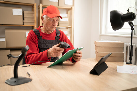 Elderly postal worker scanning barcode on parcel with handheld scanner at workplace. Preparing packages for shipment, logistics operations and mail delivery
