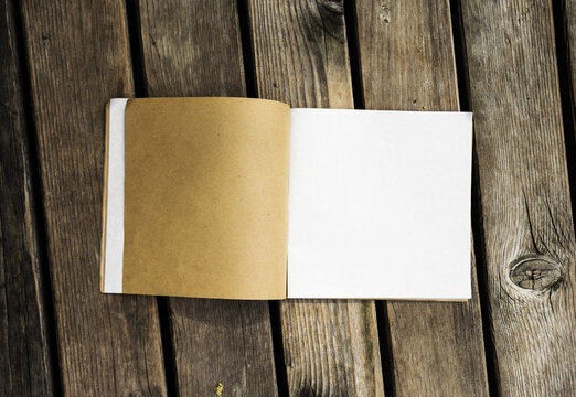 Top view of open notebook with white and kraft paper pages on a rustic wooden background. Blank book mockup with space for design.