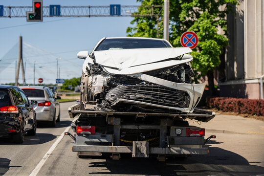 Damaged automobile with smashed front bumper carried by recovery vehicle on urban highway, van&scaron;u bridge background, vehicle evacuation and towing logistics services