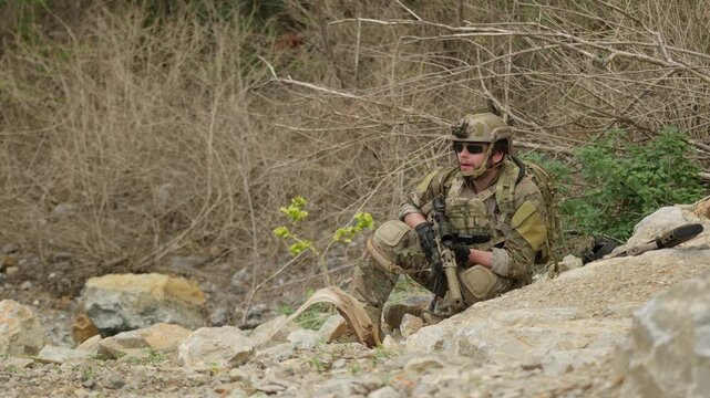 A special forces operator in full combat gear maintains a tactical overwatch position on a rugged, rocky hillside during a covert military operation.