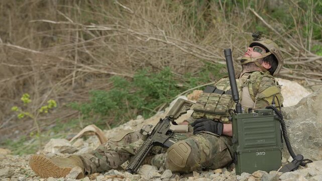 A special forces soldier rests and rehydrates in a rugged environment, awaiting orders on his tactical radio during a demanding military mission.