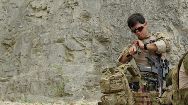 A focused military operator prepares his tactical gear, loading a magazine against a rugged cliff face to ensure operational readiness for a mission.