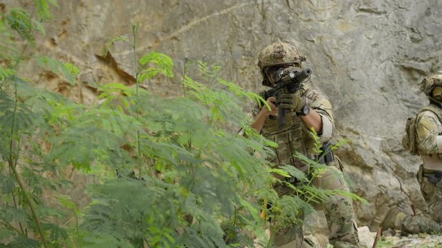 Two soldiers in full combat gear conduct a tactical patrol, securing the perimeter with assault rifles in a rugged, rocky environment during a military operation.